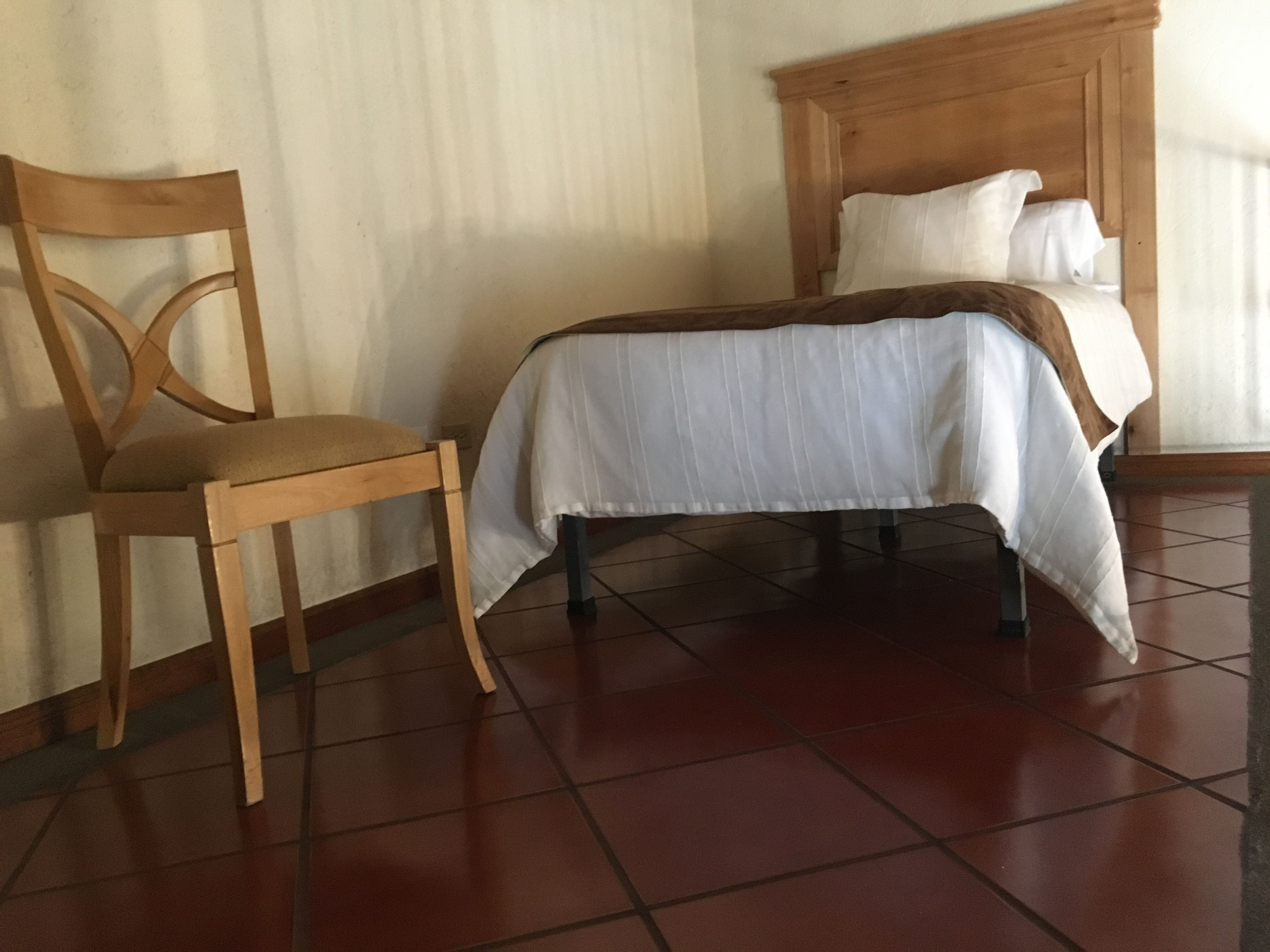 Una habitación con una cama, una silla de madera y suelo de baldosas.