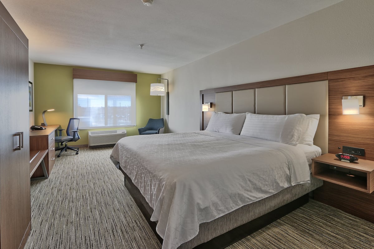 A hotel room with a large bed, a desk with a chair, a window with blinds, and a wall-mounted nightstand with a clock.