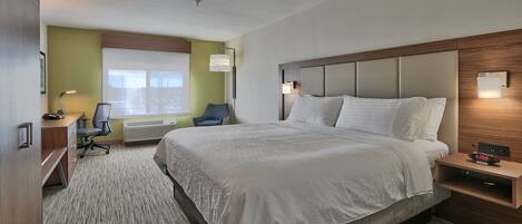 A hotel room with a large bed, a desk with a chair, a window with blinds, and a wall-mounted nightstand with a clock.