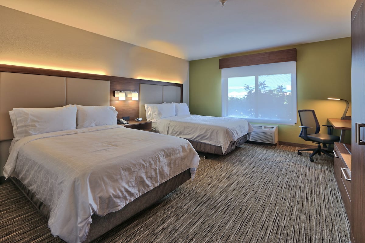 A hotel room with two beds, a desk, a chair, and a large window with a view of trees.