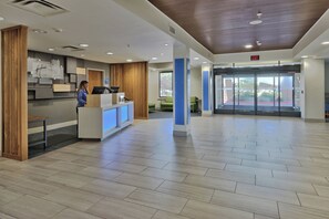 A reception area with a wooden reception desk, a woman standing behind it, and a glass door entrance. - Holiday Inn Express & Suites Albuquerque Historic Old Town by IHG (Albuquerque)