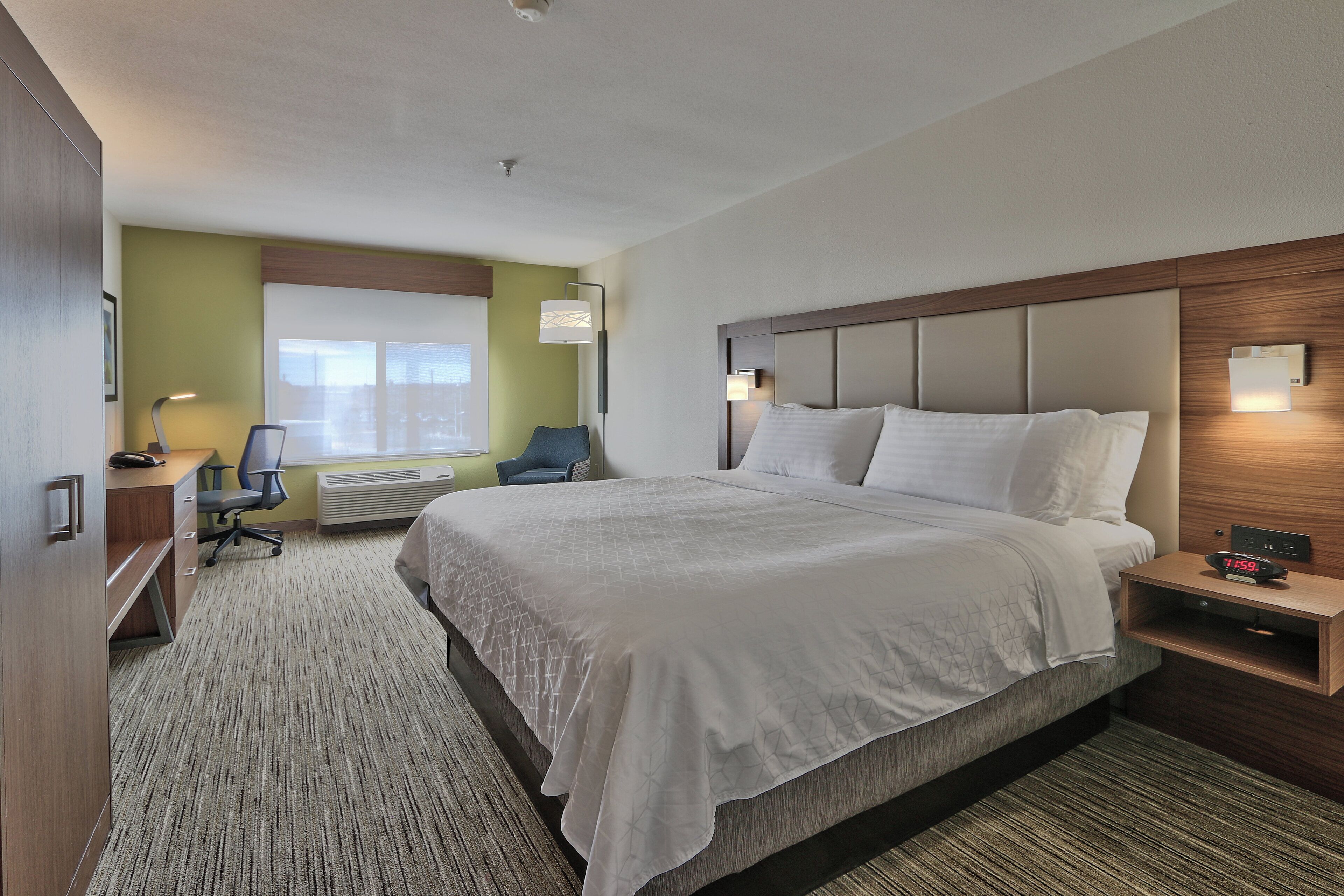 A hotel room with a large bed, a desk with a chair, a window, and a nightstand with a clock.