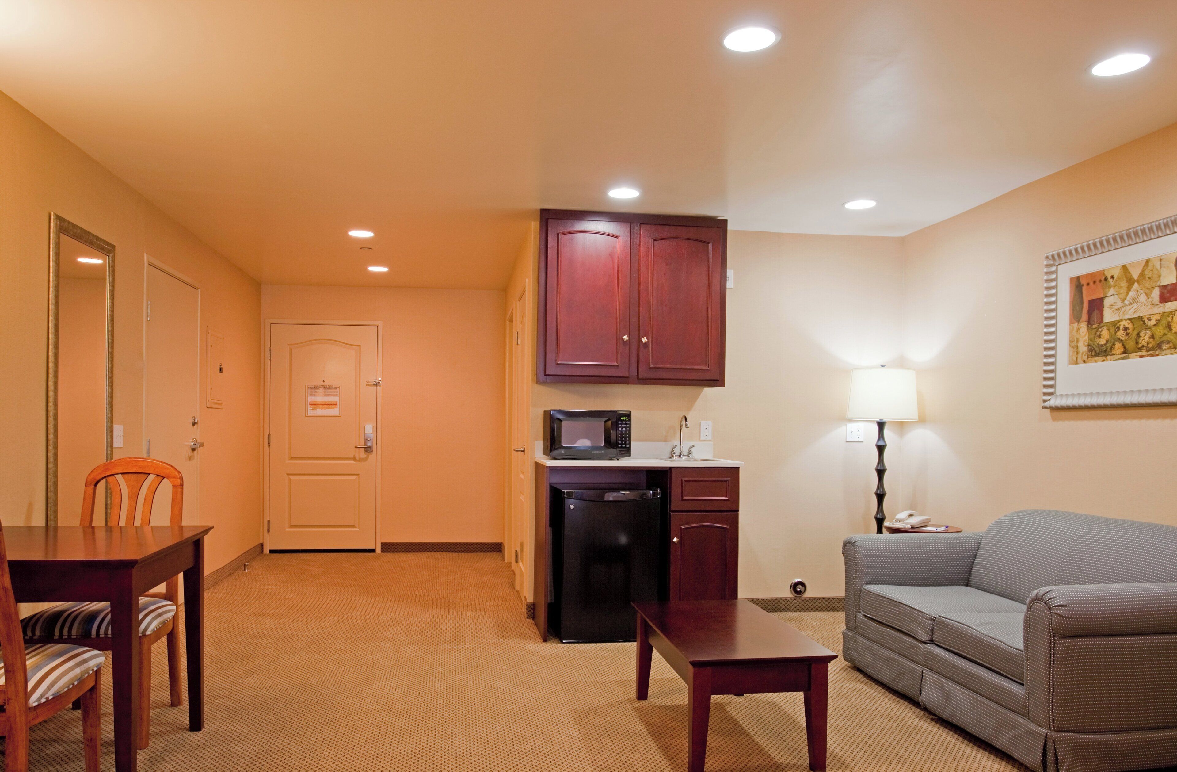 A compact hotel room with a kitchenette, a dining area with a table and chairs, a sofa, and a lamp.