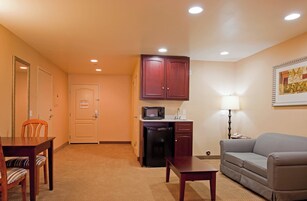A compact hotel room with a kitchenette, a dining area with a table and chairs, a sofa, and a lamp.