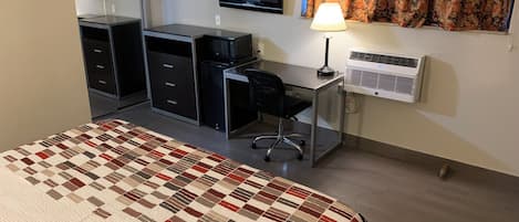 A hotel room with a bed, a desk with a chair, a television, a lamp, and an air conditioning unit.