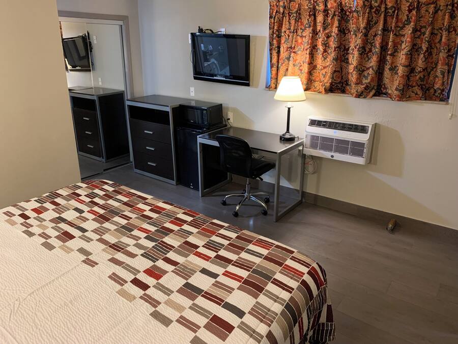 A hotel room with a bed, a desk with a chair, a television, a lamp, and an air conditioning unit.