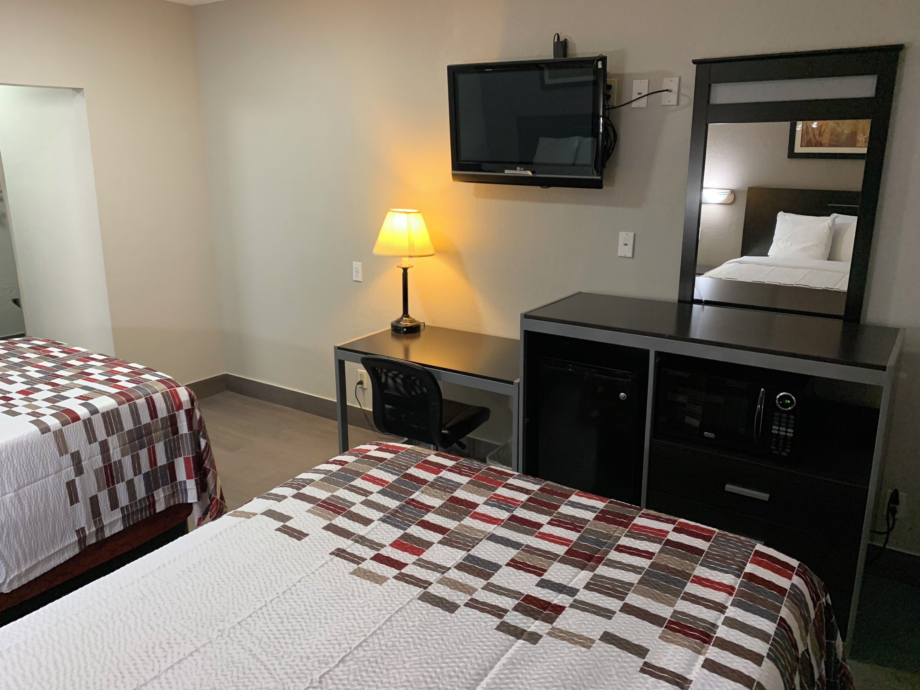 A hotel room with two beds, a desk with a lamp, a television, a microwave, and a mirror.