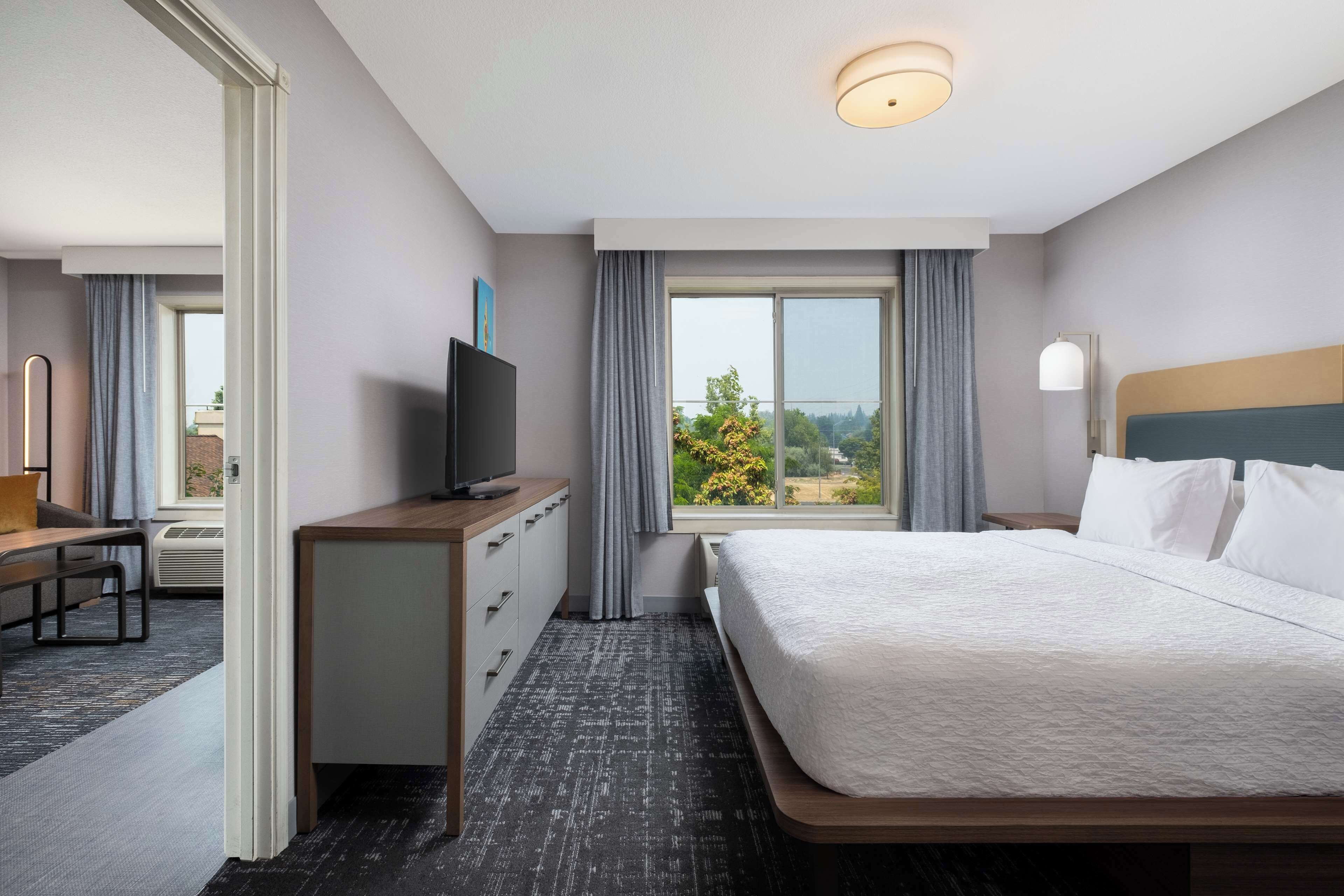 Habitación de hotel con una cama grande, un escritorio con televisor, una ventana con cortinas y vistas al exterior.