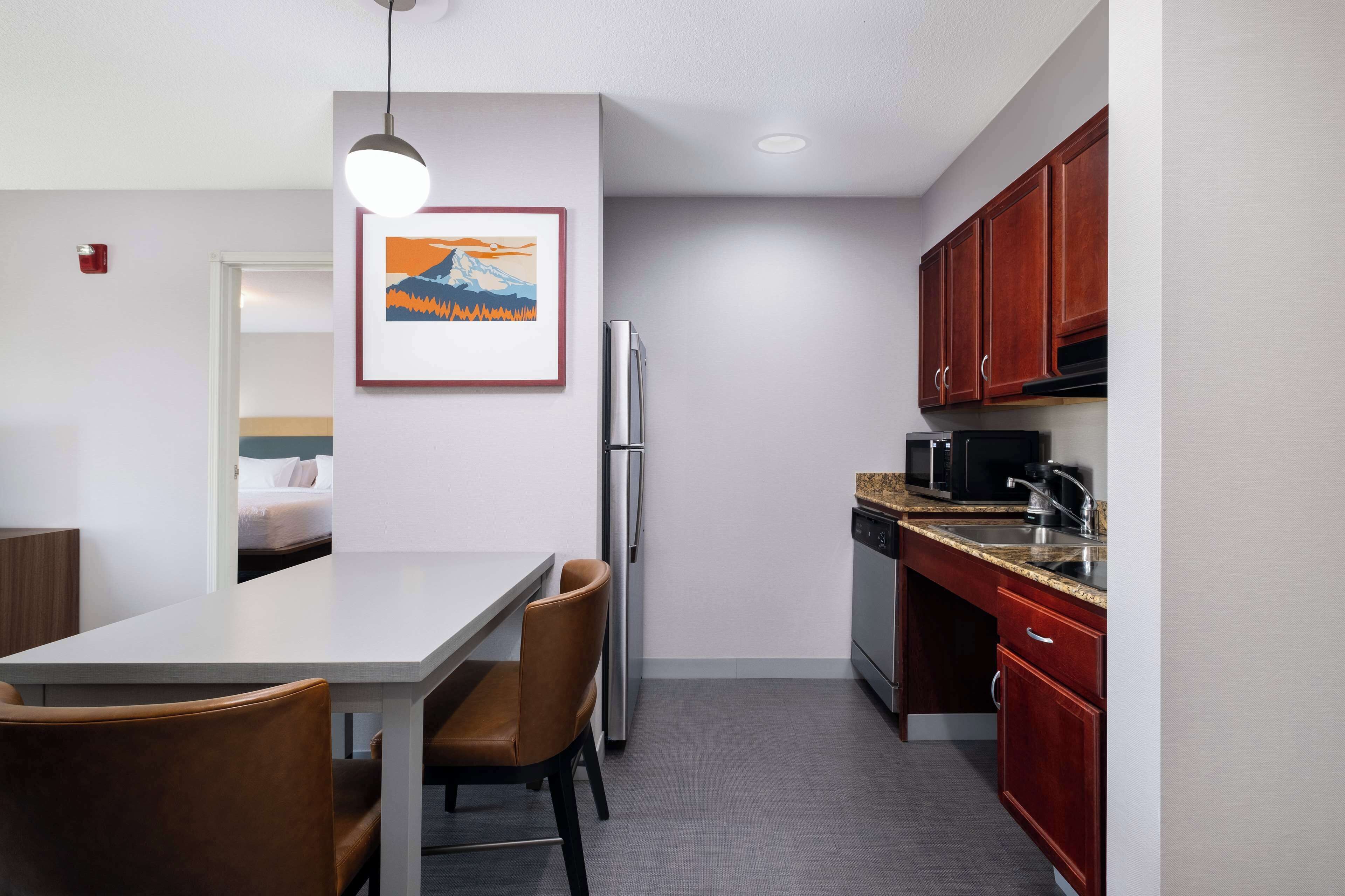 A compact kitchen with a refrigerator, microwave, and stove. There is a dining table with chairs and a pendant light. A framed picture hangs on the wall.