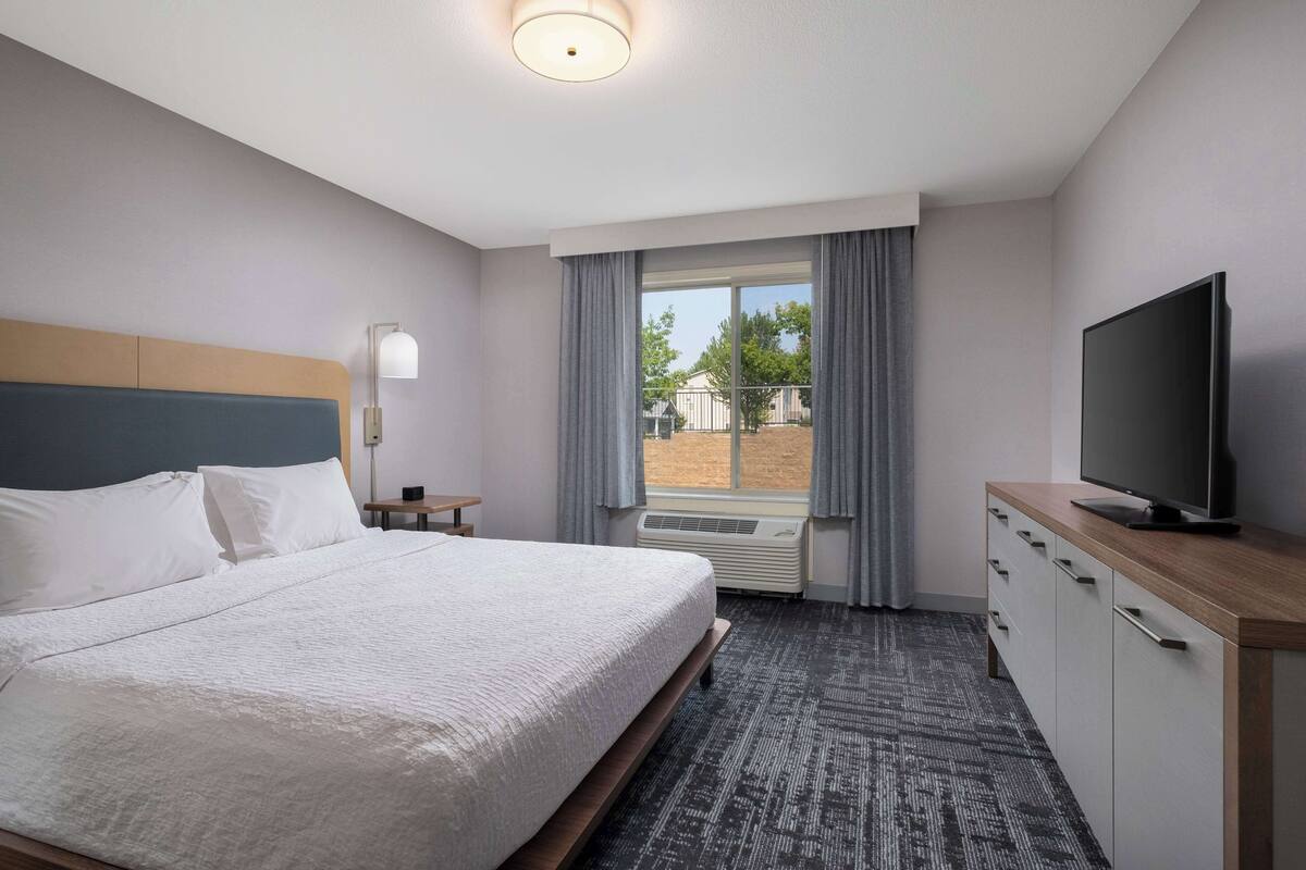 A hotel room with a large bed, a flat-screen TV, a desk, and a window with curtains.