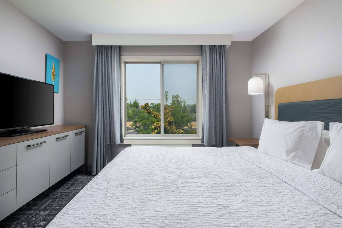 A hotel room with a large bed, a flat-screen TV, a wooden side table, and a window with curtains.