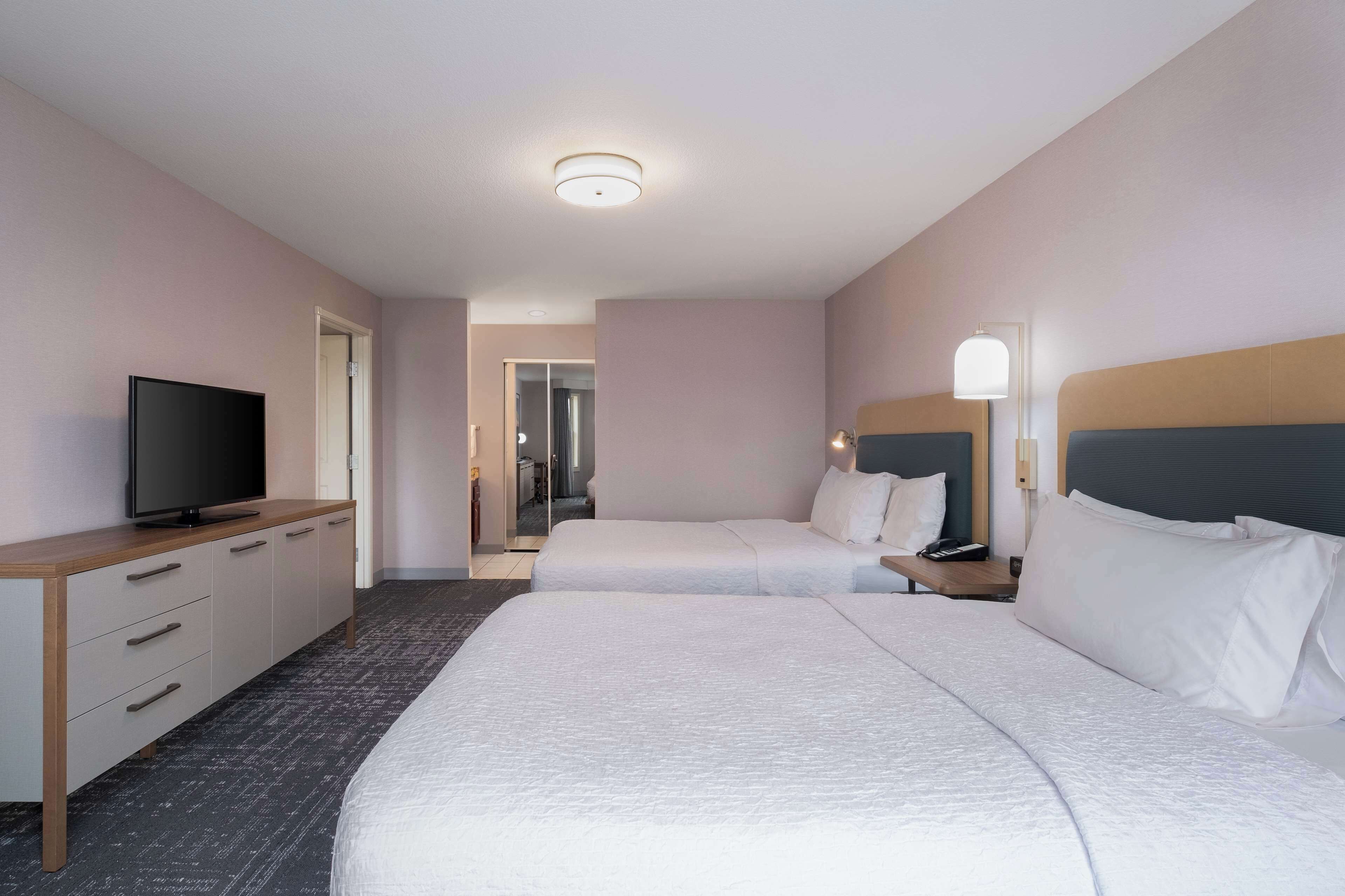 Una habitación de hotel con dos camas, un televisor sobre un aparador de madera y una mesita de noche con lámpara.