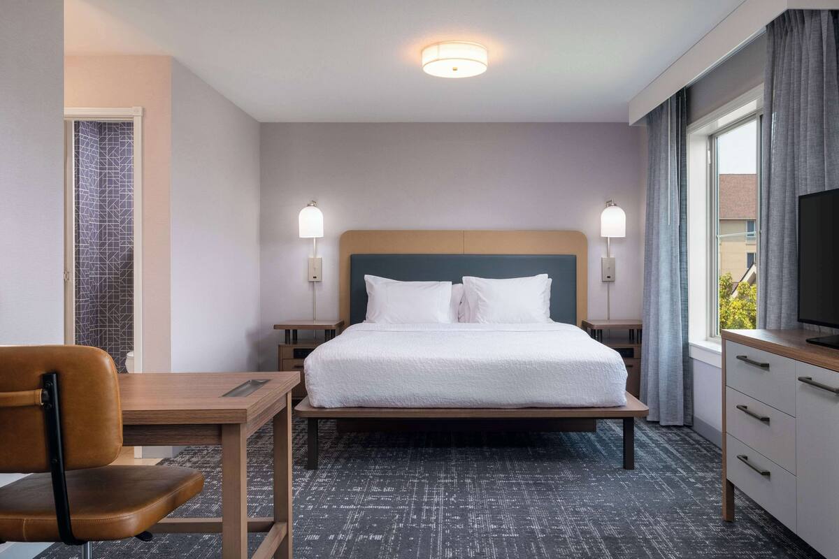 A hotel room with a bed, bedside tables, a desk, a chair, a TV, and a window with curtains.