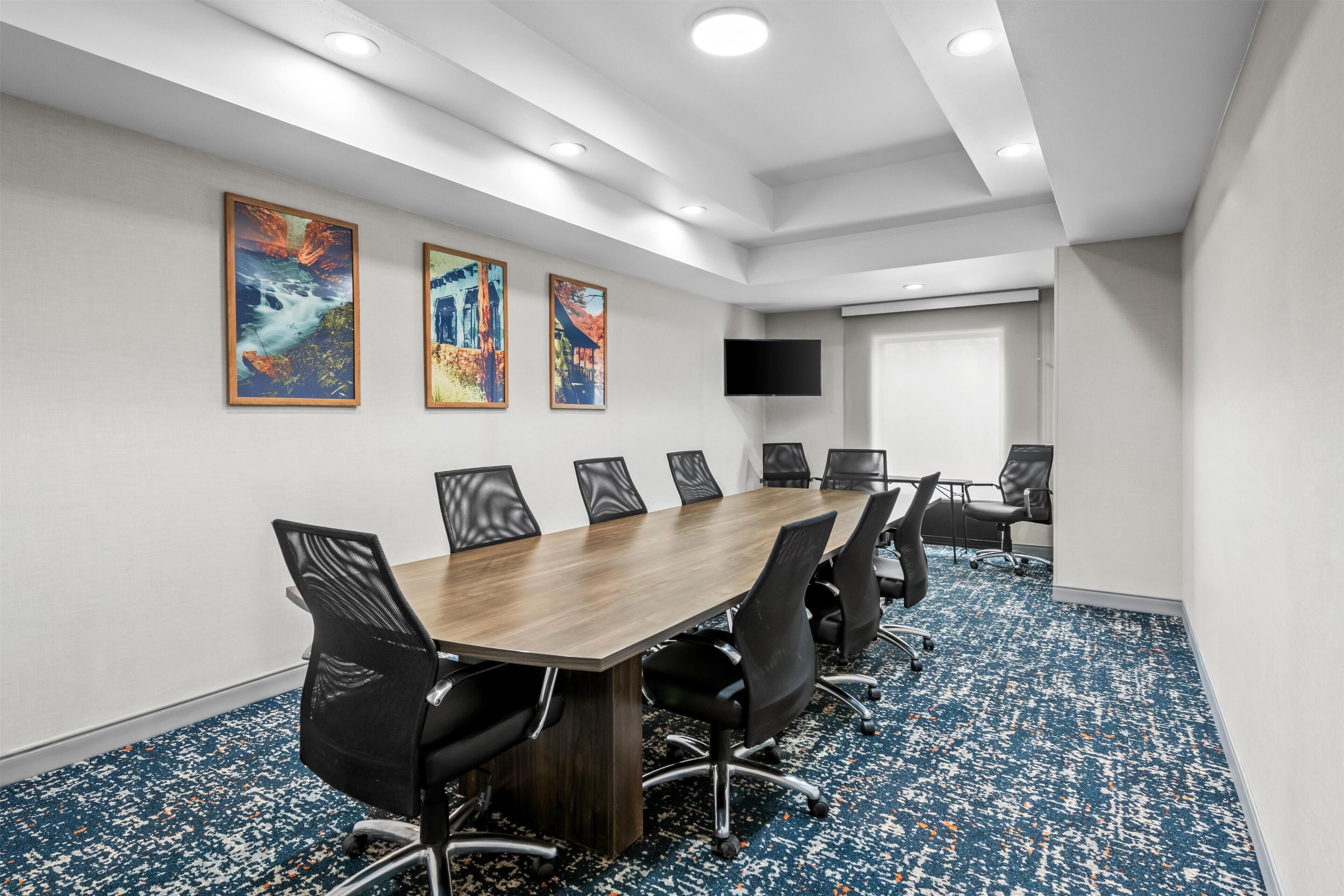 A conference room with a long table, black chairs, a flat-screen TV, and three framed artworks on the wall.