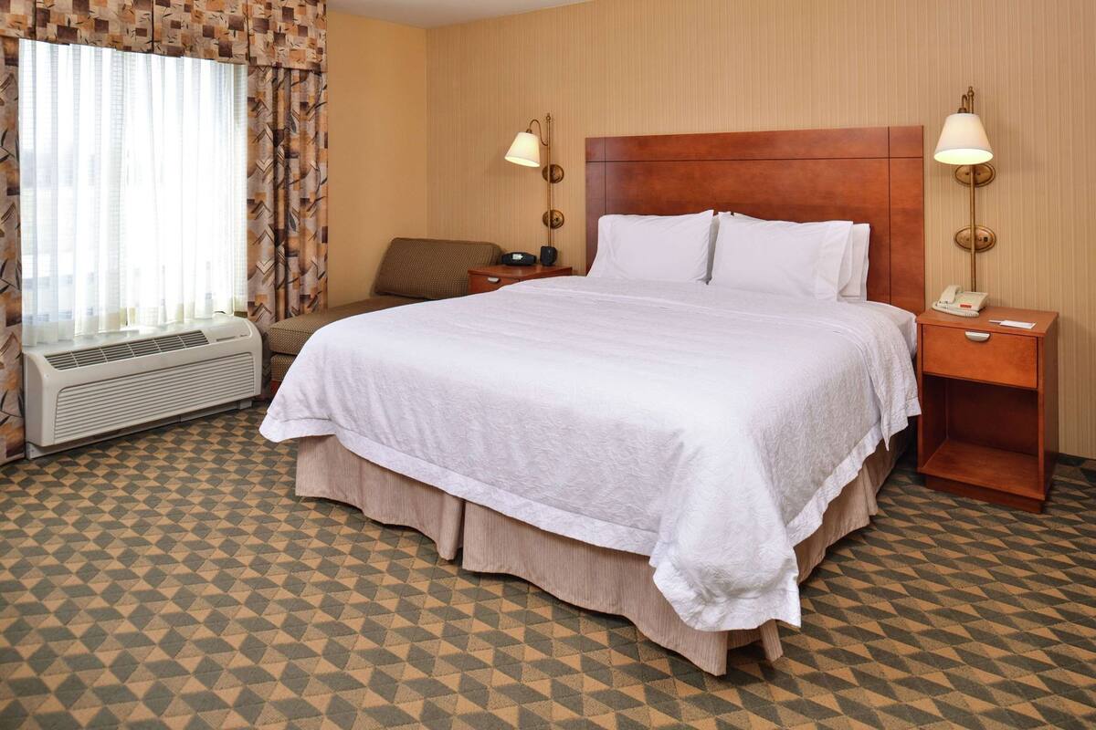 Habitación de hotel con una cama grande, dos lámparas de noche, una mesita de noche con teléfono, una silla y una ventana con cortinas.