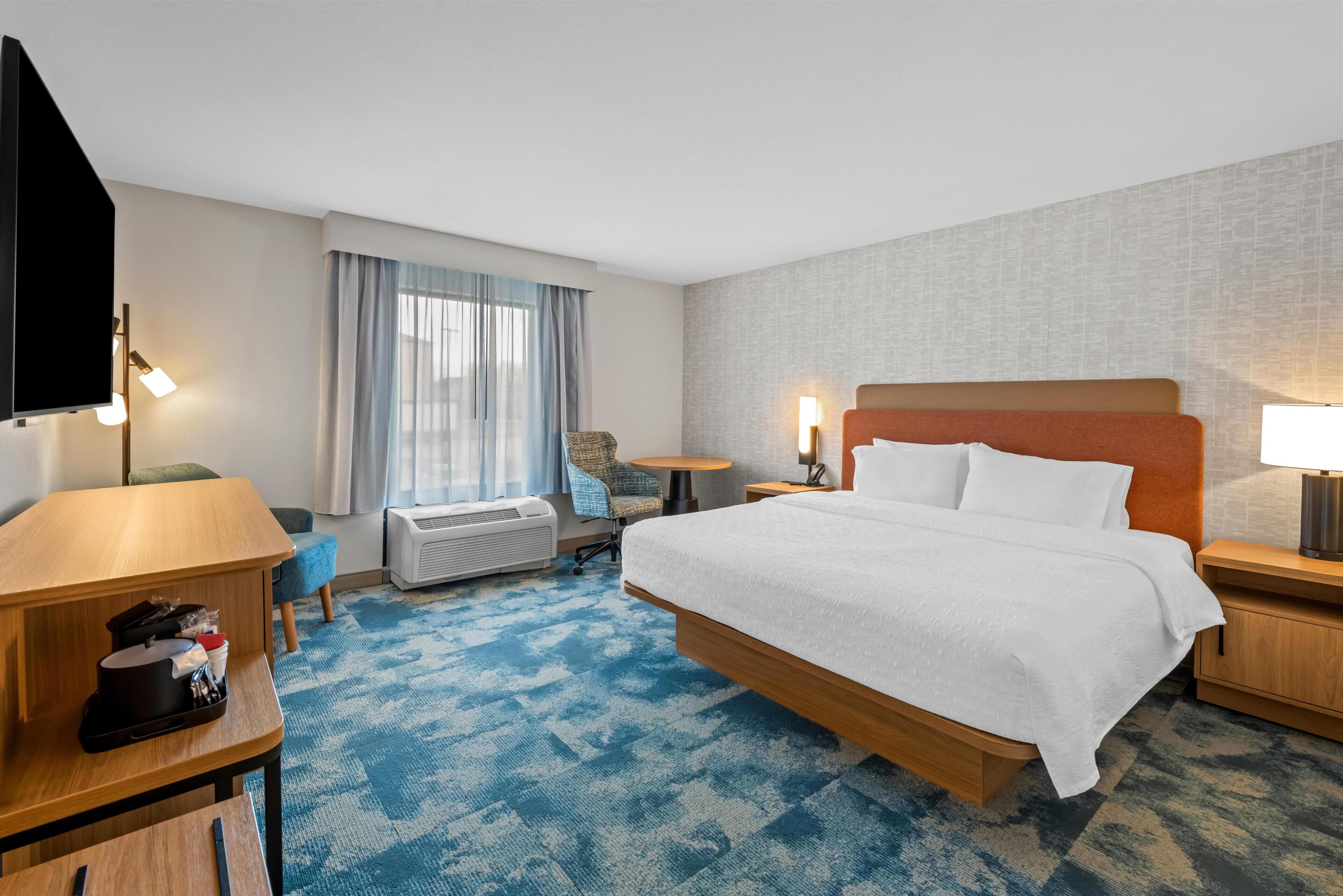 A hotel room with a large bed, a desk, a chair, a small table, a TV, and a window with curtains.