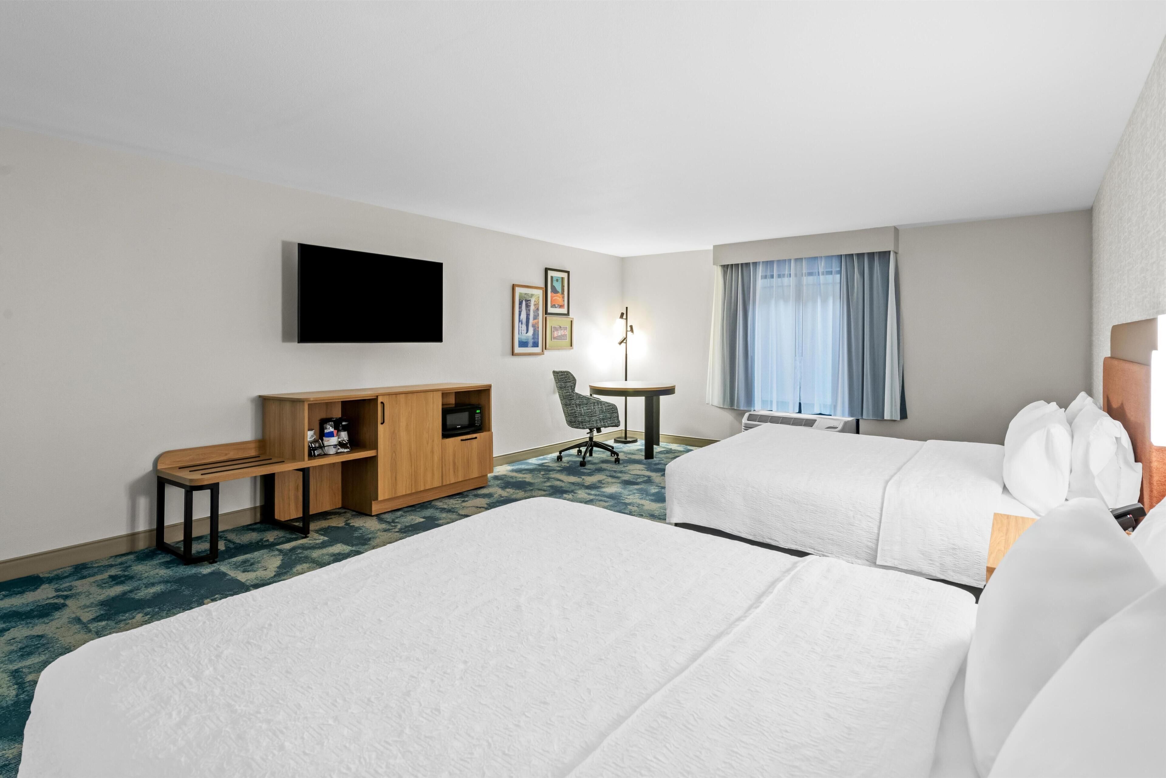 A hotel room with two beds, a desk with a microwave, a chair, a flat-screen TV, and a window with curtains.