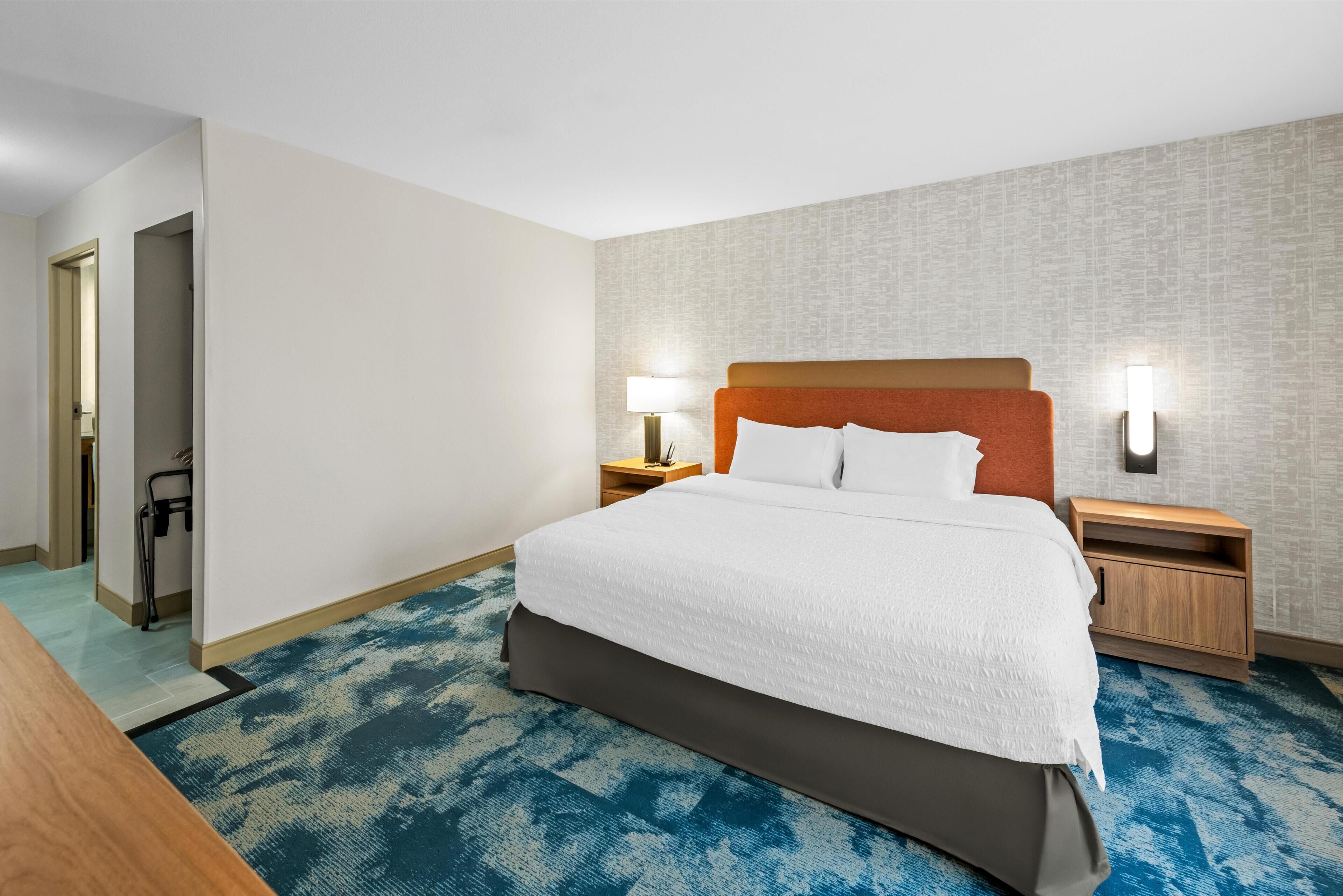 A hotel room with a large bed, two bedside tables, a nightstand, a chair, and a blue patterned carpet.