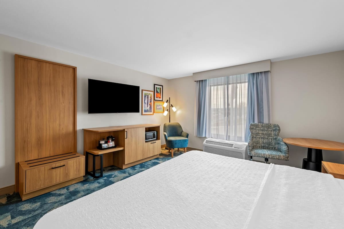 Habitación de hotel con una cama grande, un armario de madera, un escritorio con una computadora, una silla, un televisor montado en la pared y una ventana con cortinas.