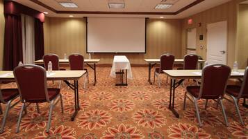 Una sala de conferencias con piso alfombrado, sillas rojas, mesas, una pantalla de proyector y una pizarra.