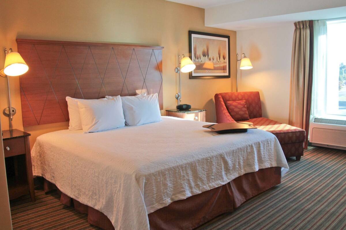 A hotel room with a large bed, two bedside lamps, a chair, a painting on the wall, and a window with curtains.