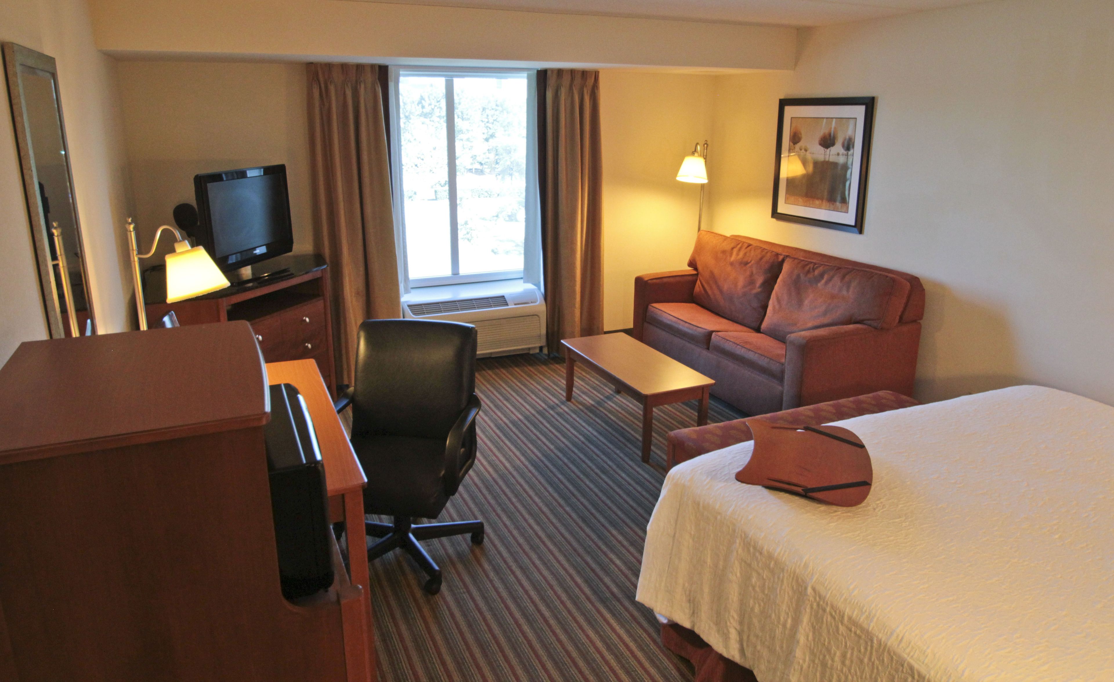 Une chambre d’hôtel avec un lit, un canapé, un bureau avec une télévision, une chaise et une fenêtre avec des rideaux.