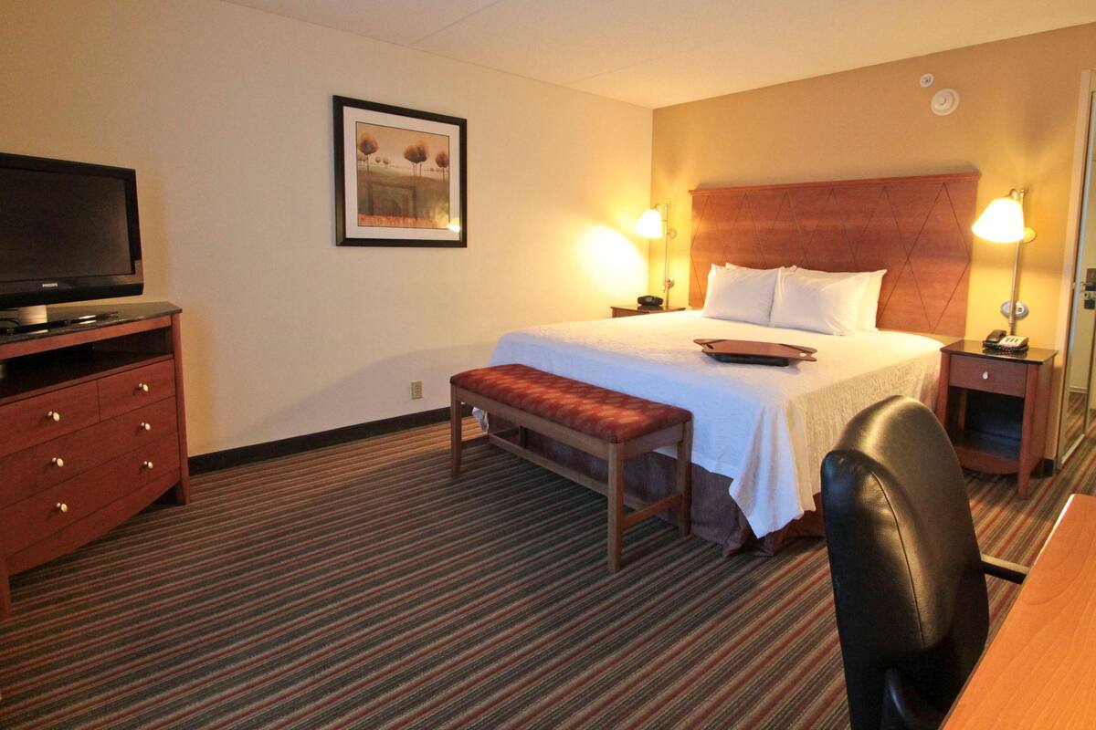 A hotel room with a bed, bedside lamps, a TV, a desk, and a bench.