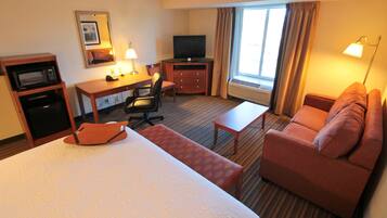 A hotel room with a bed, a desk with a television, a microwave, a chair, a sofa, and a window with curtains.
