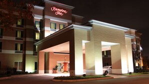 Un hotel Hampton Inn con una entrada bien iluminada, un estacionamiento visible y una vista clara de la fachada del edificio.