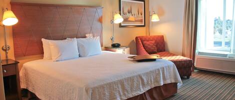 A hotel room with a large bed, two bedside lamps, a chair, a small table, and a window with curtains.
