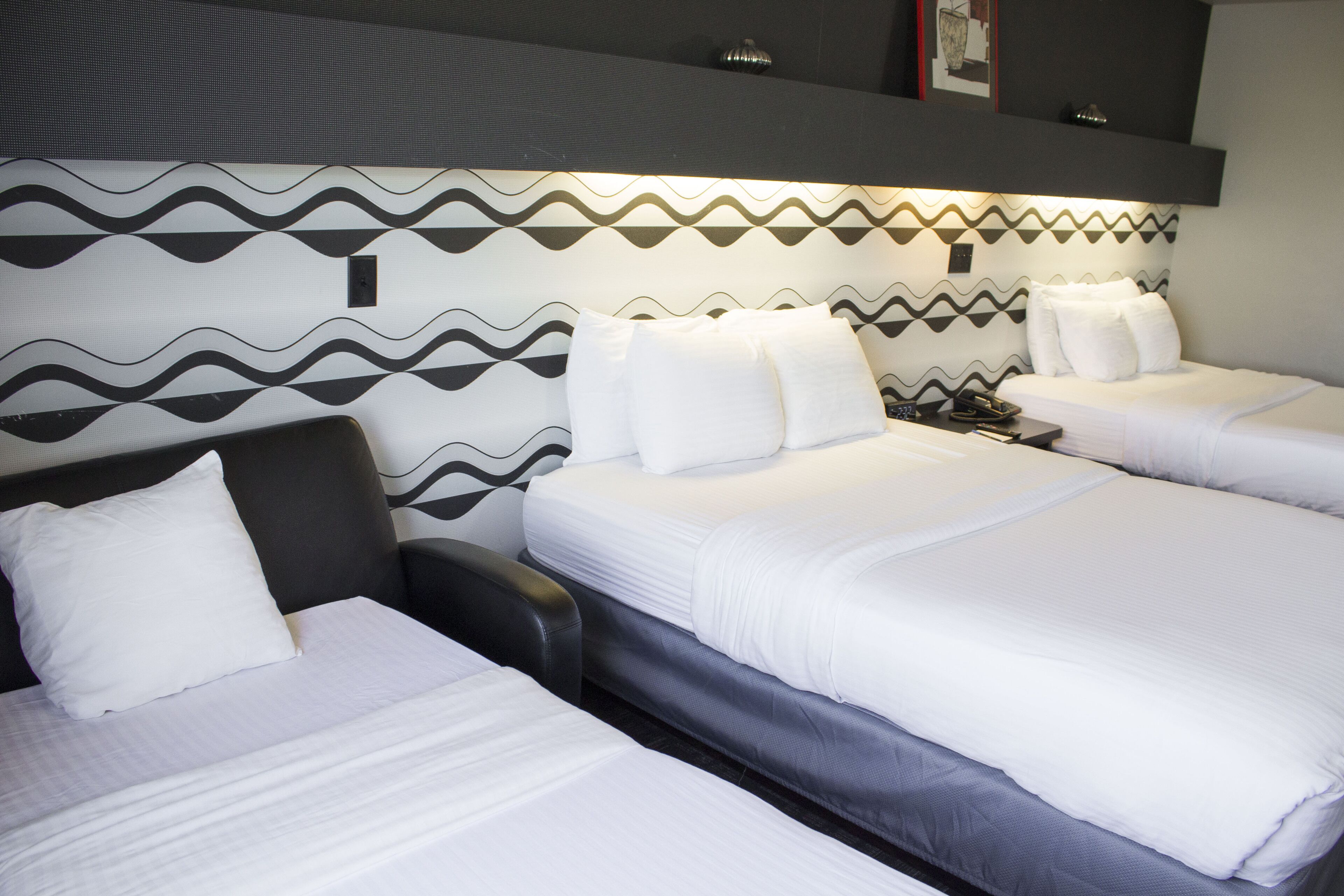 A hotel room with two beds, each with white linens and pillows. The headboard has a geometric pattern, and there is a small table with a lamp between the beds.