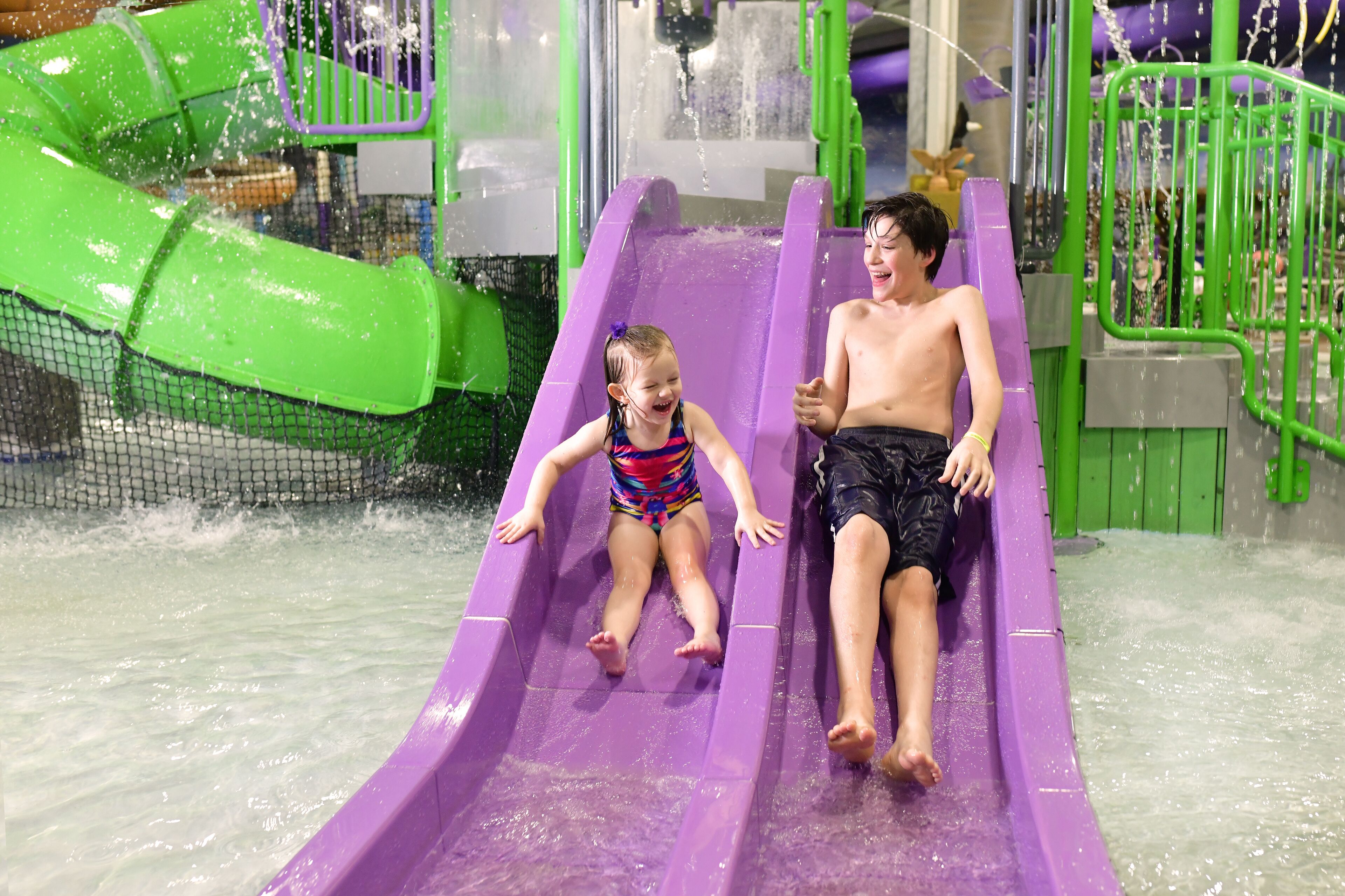 Two children sliding down a purple water slide in an indoor water park.