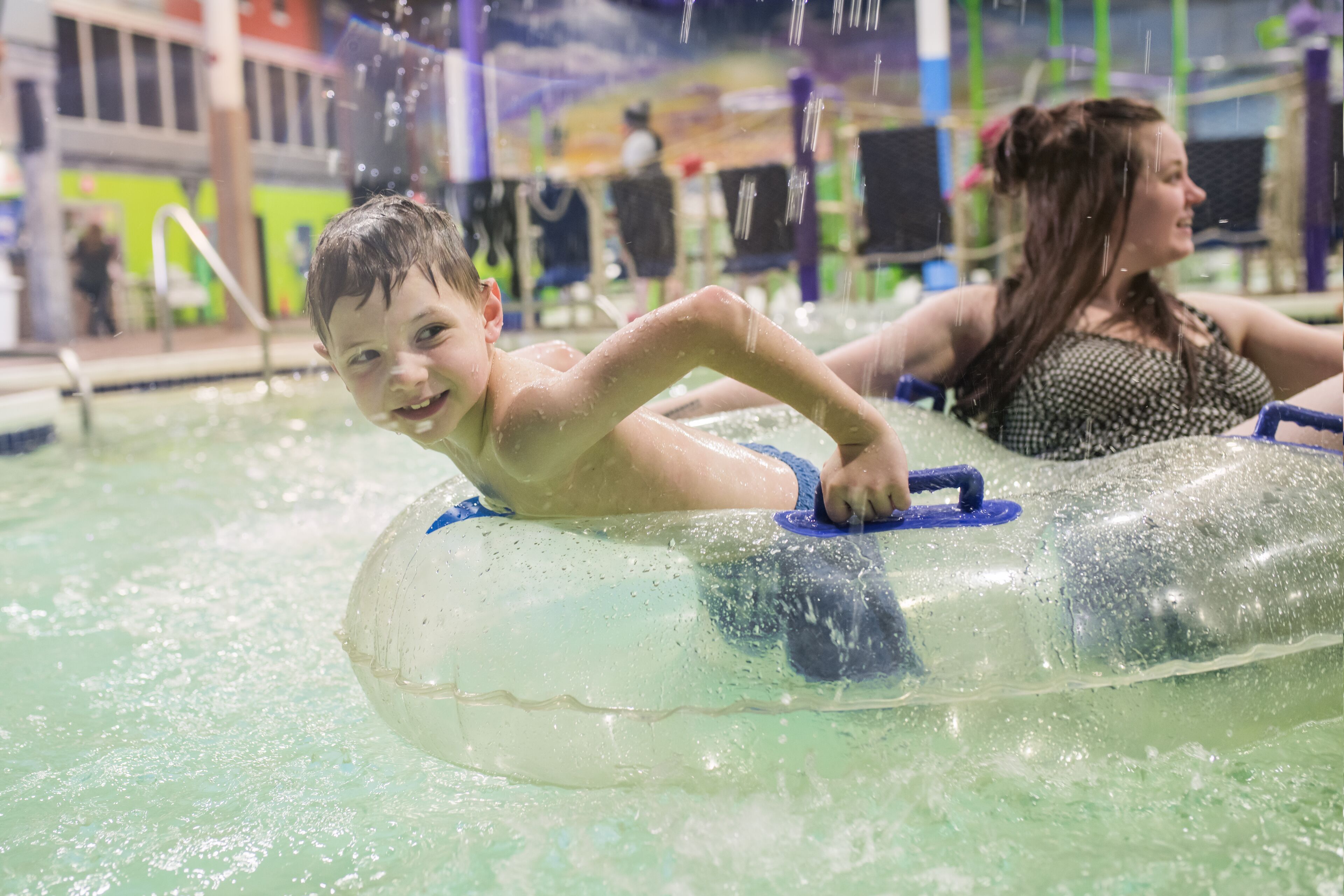 A child and an adult in an indoor water park, floating on a clear water tube.