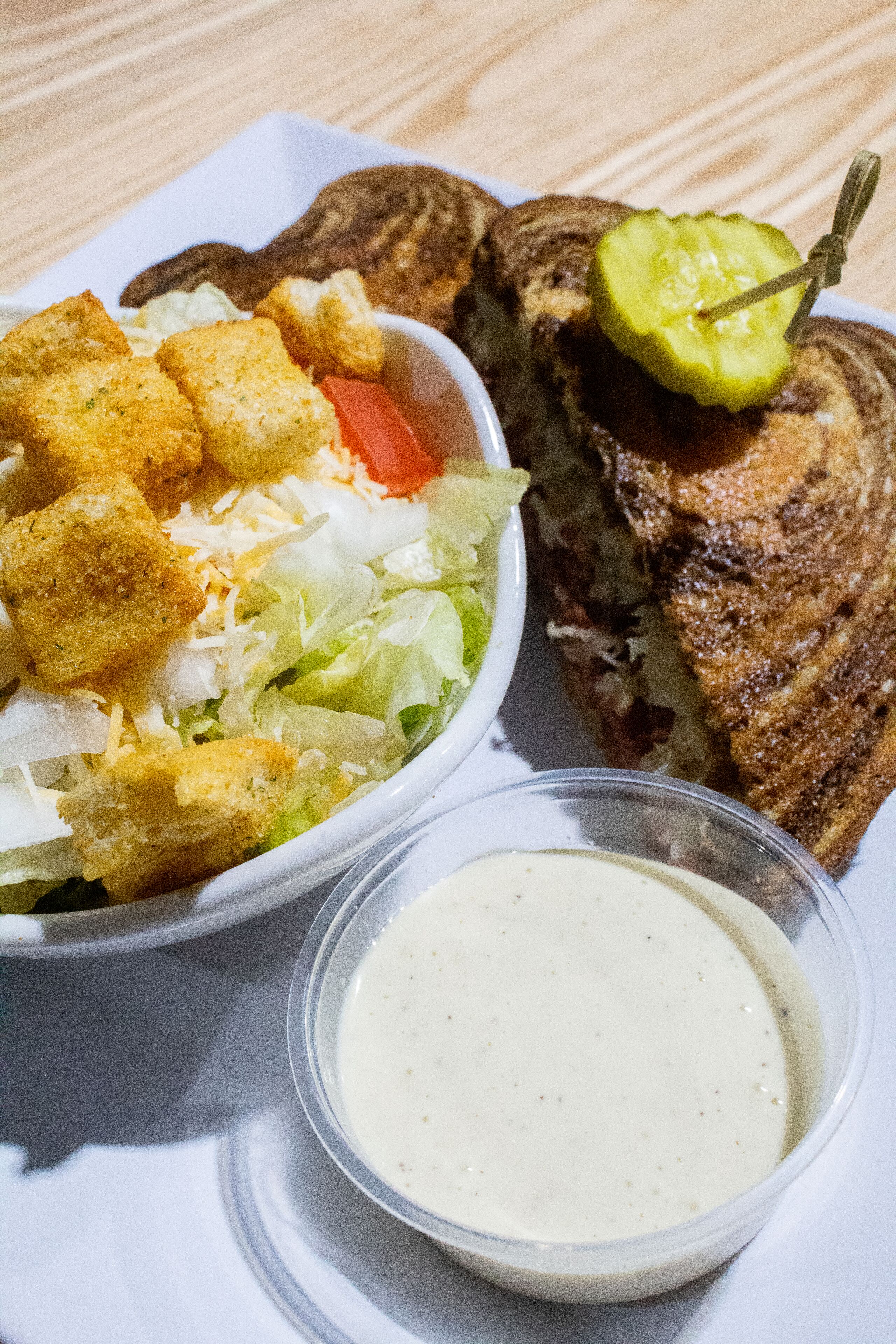 A sandwich with a pickle on top, a side salad with croutons, and a small container of sauce.