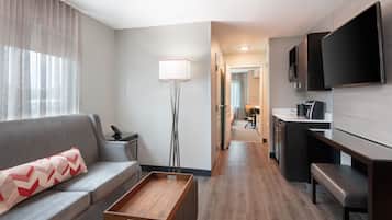 A modern hotel room with a sofa, a coffee table, a floor lamp, a TV, and a kitchenette.