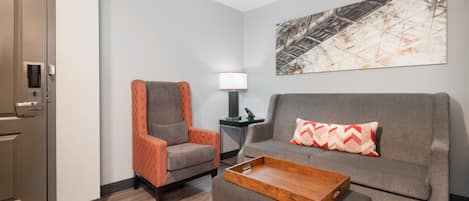 A hotel room with a grey sofa, an orange armchair, a wooden tray table, a lamp, and a large abstract painting on the wall.