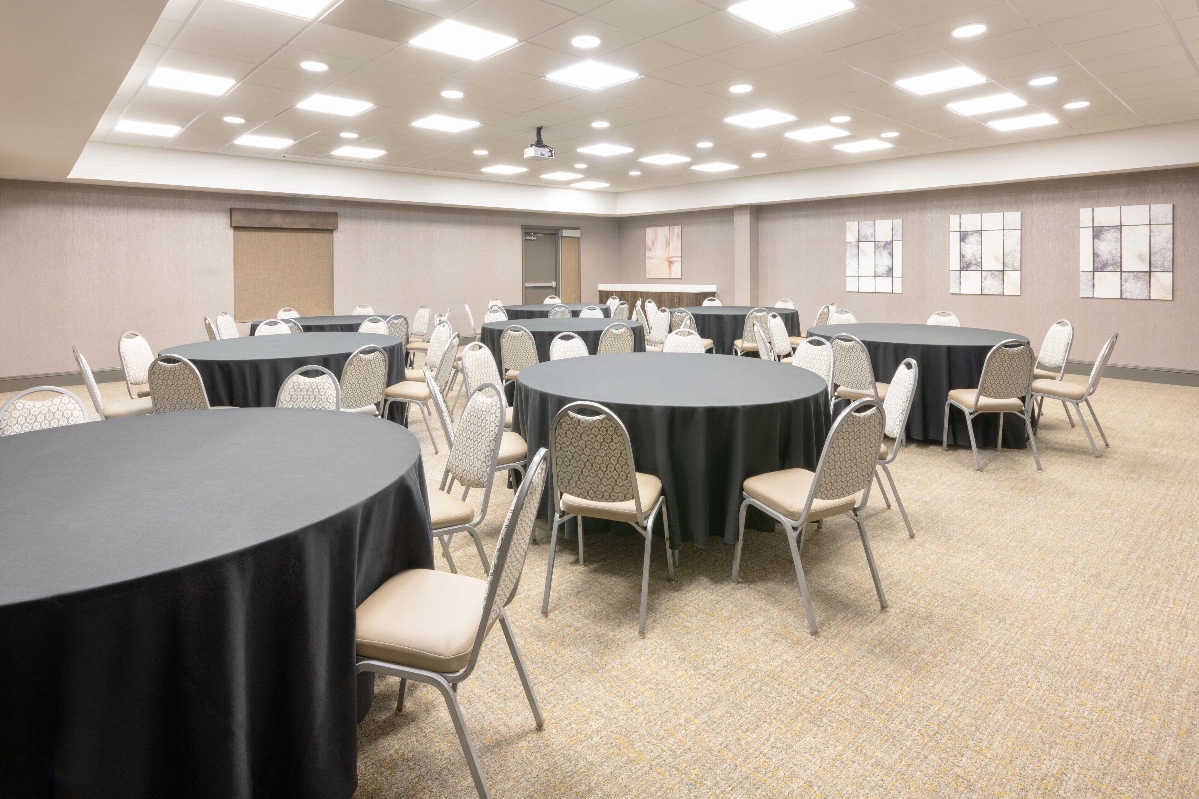 A conference room with multiple round tables, each surrounded by chairs. The room has a carpeted floor and a high ceiling with recessed lighting.