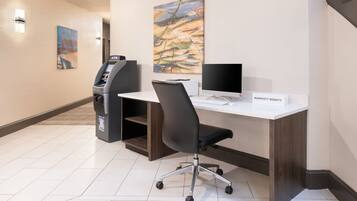 A hotel workspace with a desk, computer, chair, and ATM.