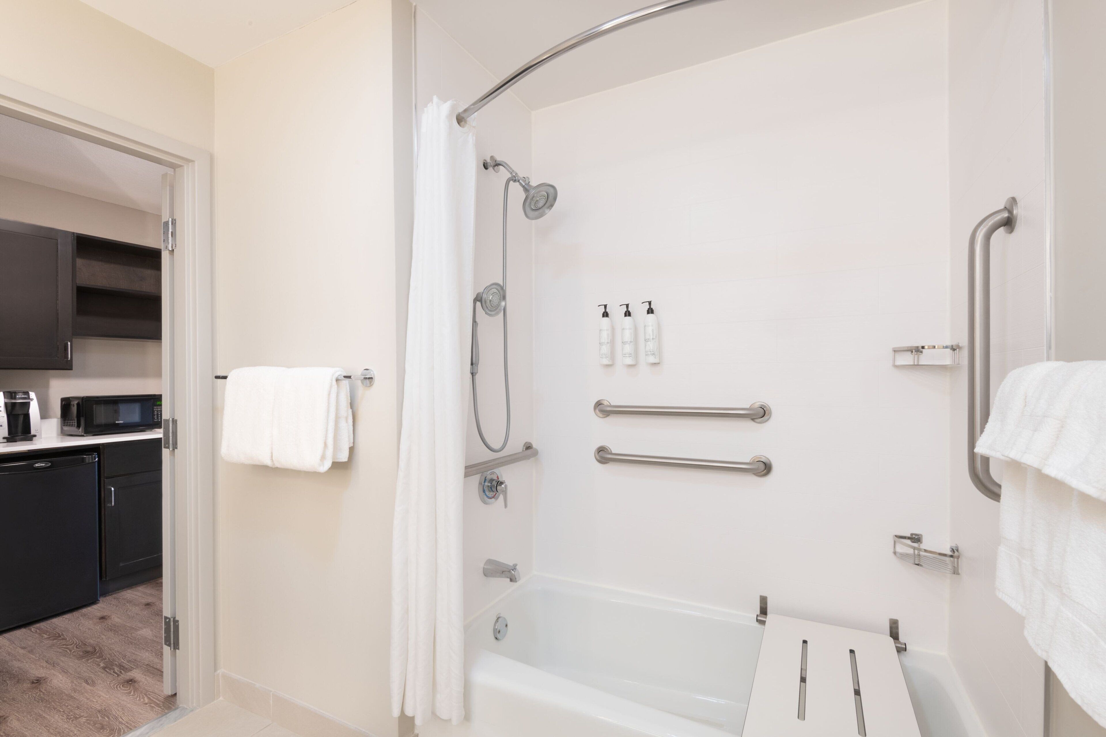 Un baño moderno con bañera, ducha y barras de apoyo. Hay artículos de aseo en la pared y un perchero para toallas.