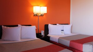 A hotel room with two beds, white bedding, a nightstand with a lamp, and an orange wall. - Motel 6 Goodland, KS (Goodland)