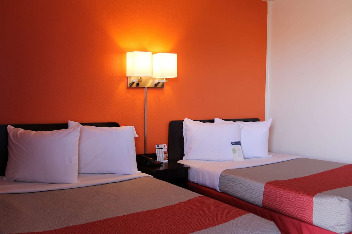 A hotel room with two beds, white bedding, a nightstand with a lamp, and an orange wall.