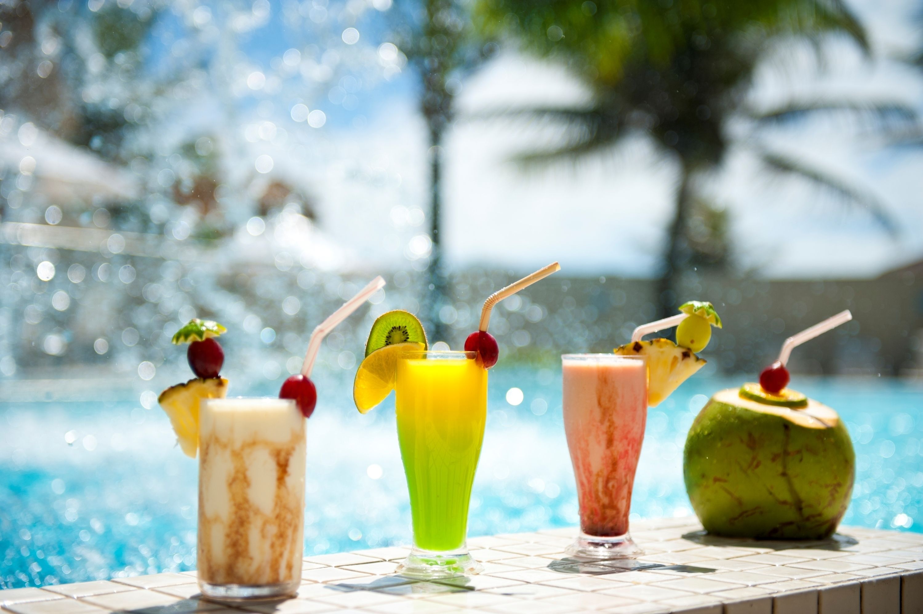 Un bar au bord de la piscine proposant une variété de boissons colorées, notamment un cocktail à l’ananas, une boisson verte, une boisson rose et une boisson à la noix de coco, avec une piscine et des palmiers en arrière-plan.