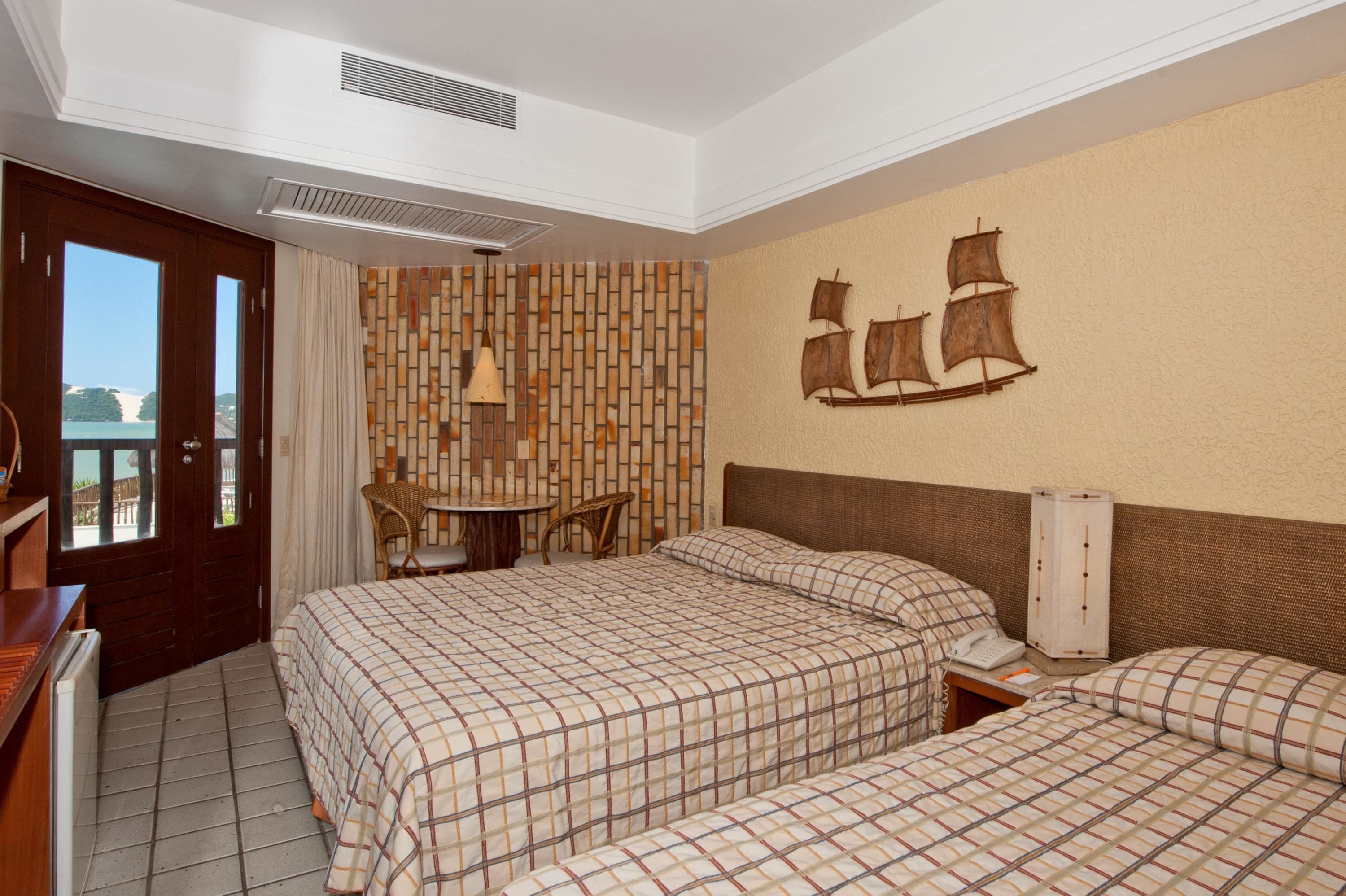 A hotel room with two beds, a small table, chairs, a view of the sea, and a ship decoration on the wall.