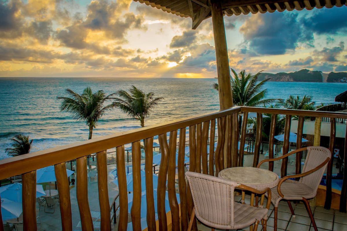 A wooden balcony with wicker chairs overlooking a beach with palm trees and a sunset.