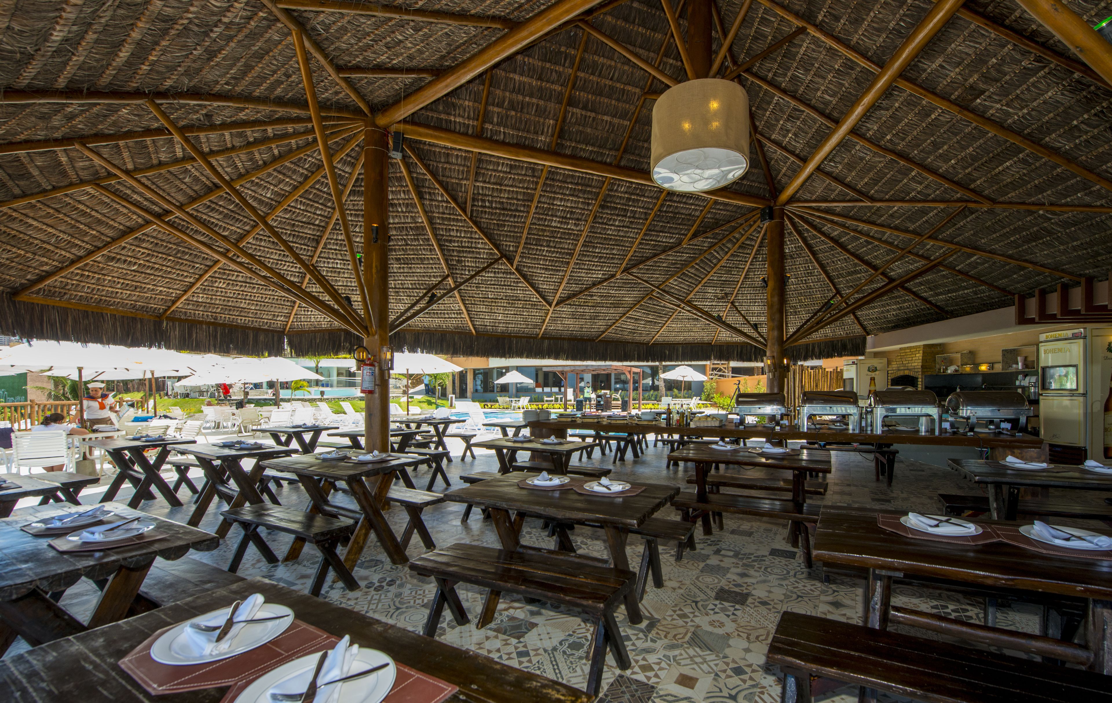 Un área de comedor al aire libre con mesas y bancos de madera, techo de paja y vista a la playa.