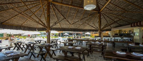 Un área de comedor al aire libre con mesas y bancos de madera, techo de paja y vista a la playa.