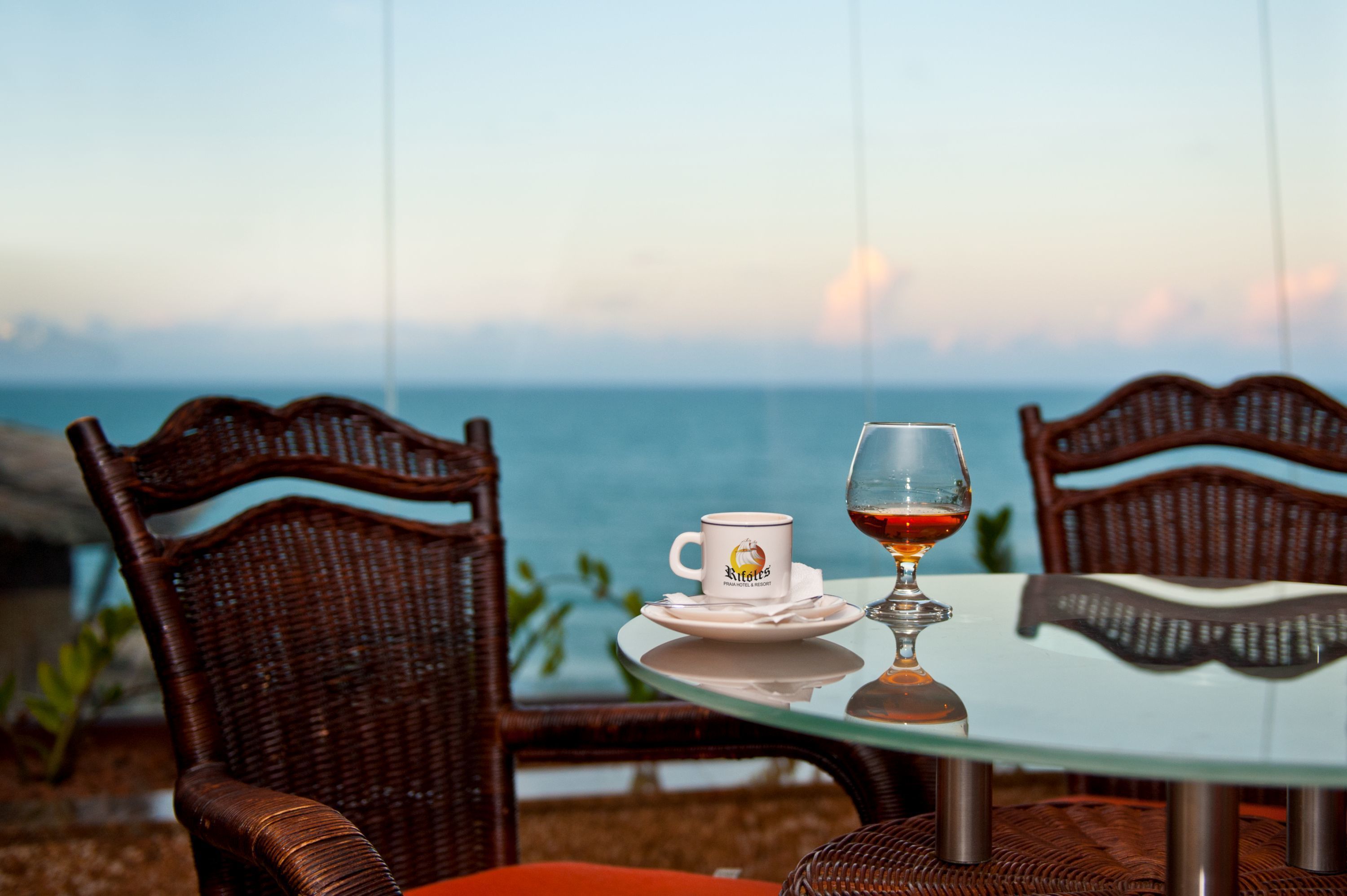 A table with a glass of red wine and a mug, wicker chairs, and a view of the sea.