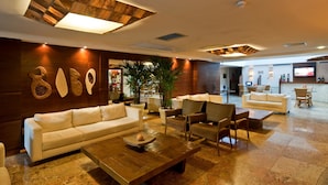 A spacious lobby with a white sofa, wooden coffee table, and several chairs. There are potted plants and a television screen on the wall.