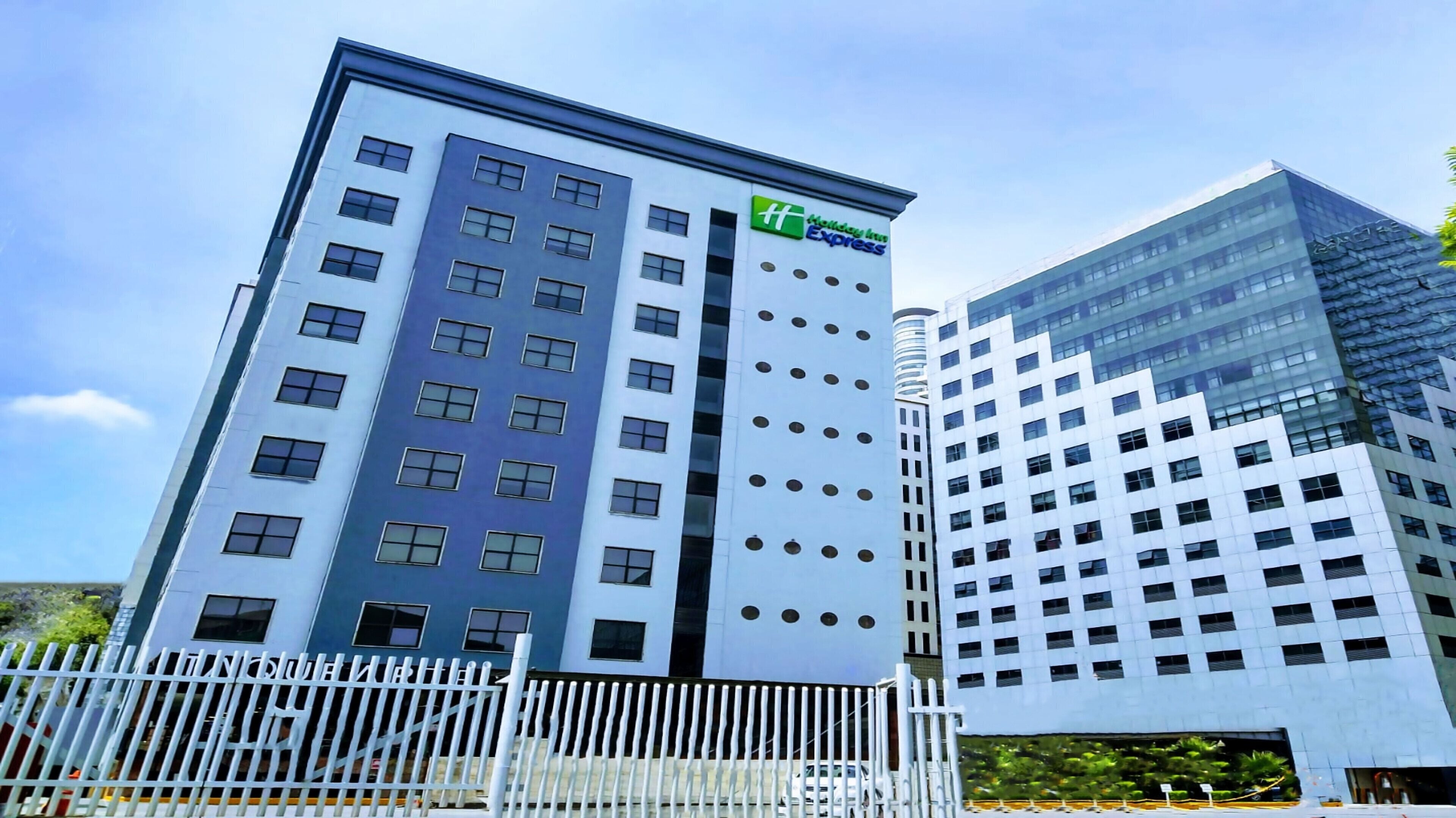 Ein modernes Hotelgebäude mit einem Holiday Inn Express Schild, das eine mehrstöckige Struktur mit einer Mischung aus rechteckigen und runden Fenstern sowie einem weißen Zaun im Vordergrund zeigt.