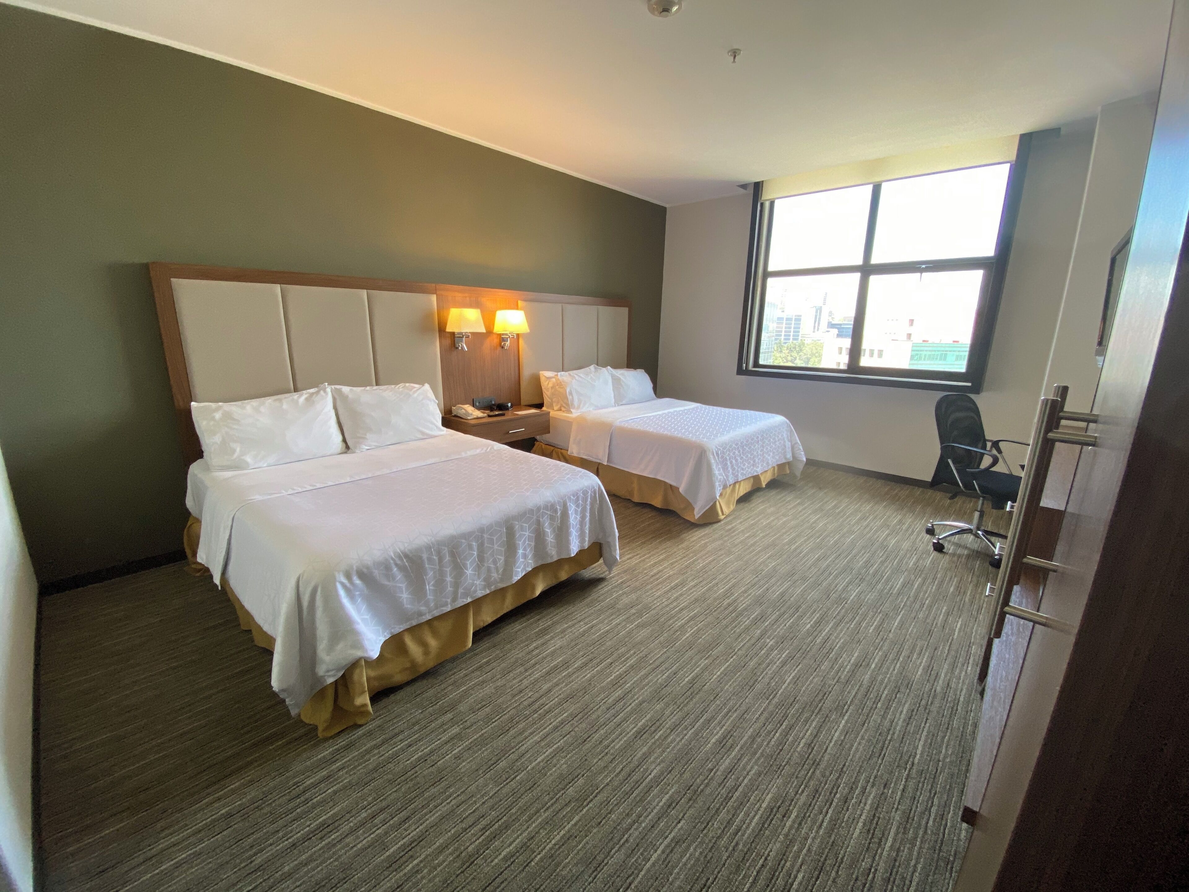 Habitación de hotel con dos camas, un escritorio, una silla, un ventanal y vista a la ciudad.